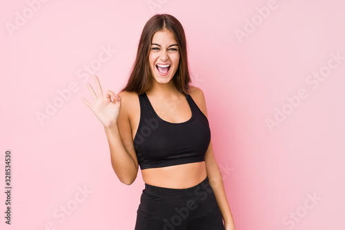 Young caucasian fitness woman doing sport isolated winks an eye and holds an okay gesture with hand.