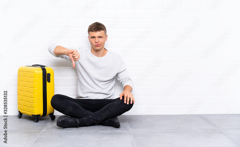 Young handsome man sitting on the floor with a suitcase showing thumb down sign Young handsome man sitting on the floor with a suitcase showing thumb down sign