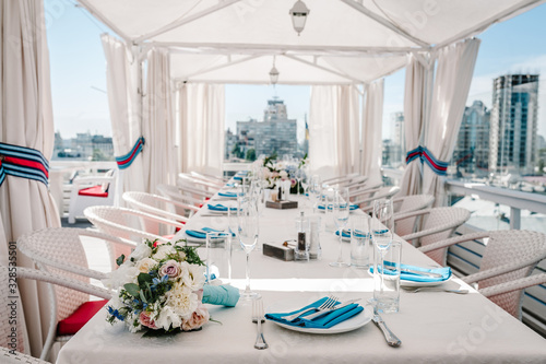 A rooftop restaurant overlooking the city. Festive table served dishes and decorated with flowers for wedding party. Image of celebrate outdoor party on background town. © Serhii