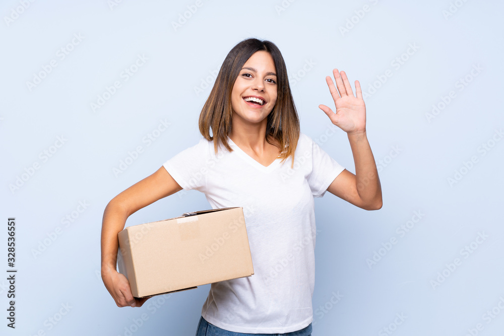 Young woman over isolated blue background holding a box to move it to another site and saluting Young woman over isolated blue background holding a box to move it to another site and saluting