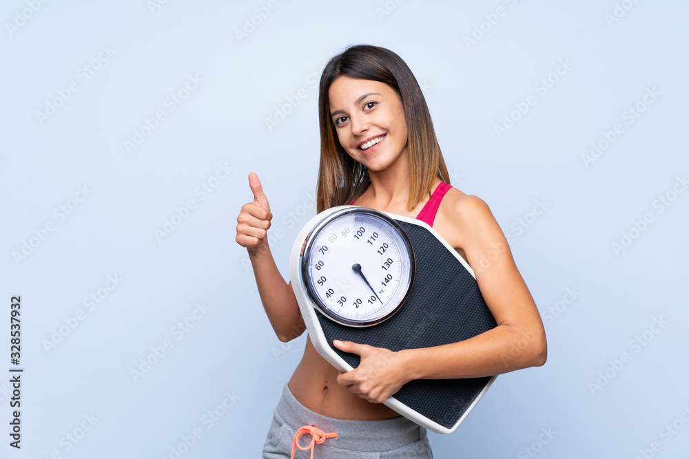 Girl with weighing machine over isolated blue background holding weighing machine with thumb up Girl with weighing machine over isolated blue background holding weighing machine with thumb up