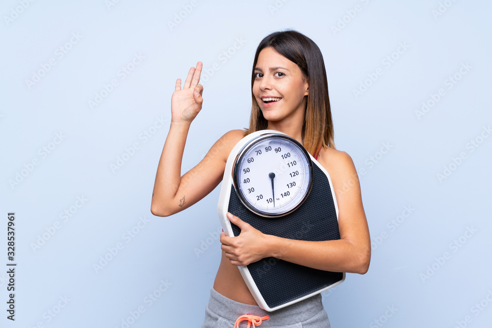 Girl with weighing machine over isolated blue background holding a weighing machine and doing OK sign Girl with weighing machine over isolated blue background holding a weighing machine and doing OK sign