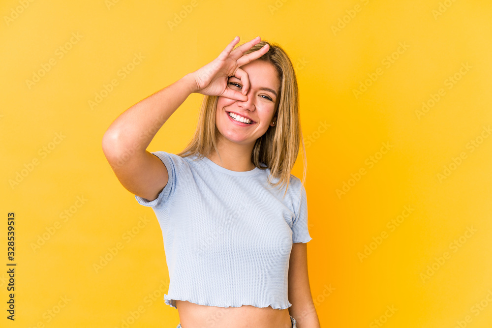 Young blonde caucasian woman isolated on yellow background excited keeping ok gesture on eye. Young blonde caucasian woman isolated on yellow background excited keeping ok gesture on eye.