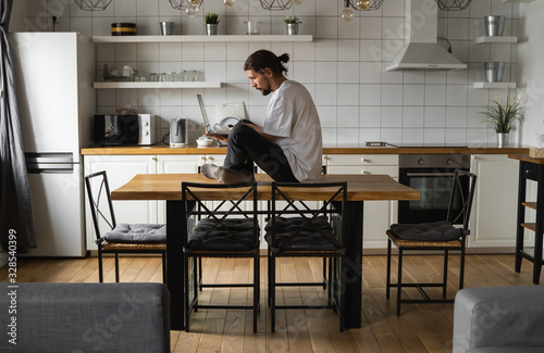 Freelancer working from home sitting on a top of the kitchen table and using laptop. Bearded man working with a laptop and reading news. Handsome successful self entrepreneur working at his modern