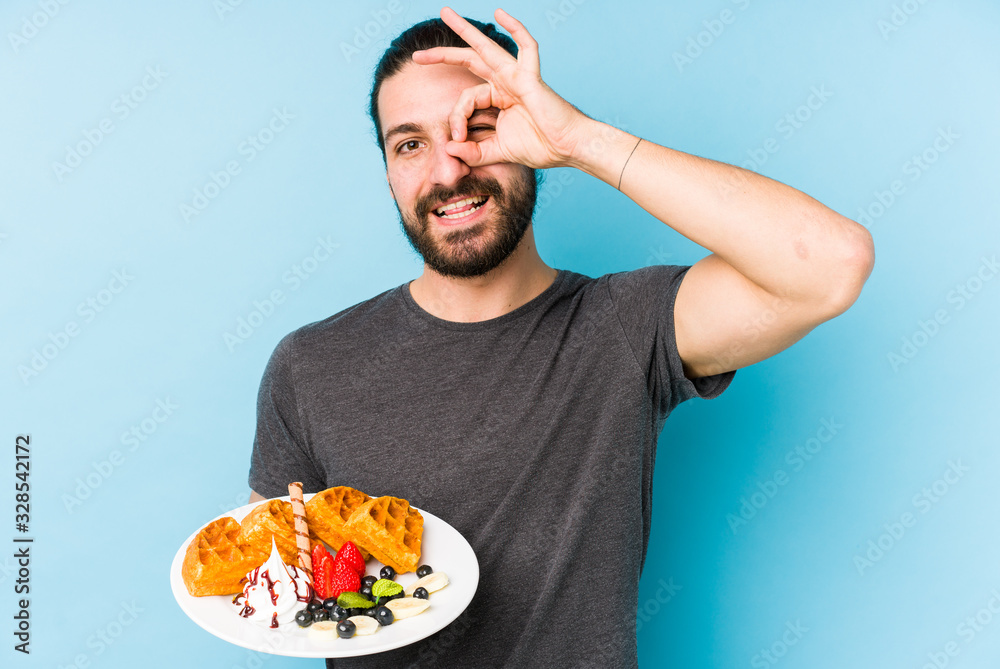 Young caucasian man eating a waffle dessert isolated excited keeping ok gesture on eye. Young caucasian man eating a waffle dessert isolated excited keeping ok gesture on eye.