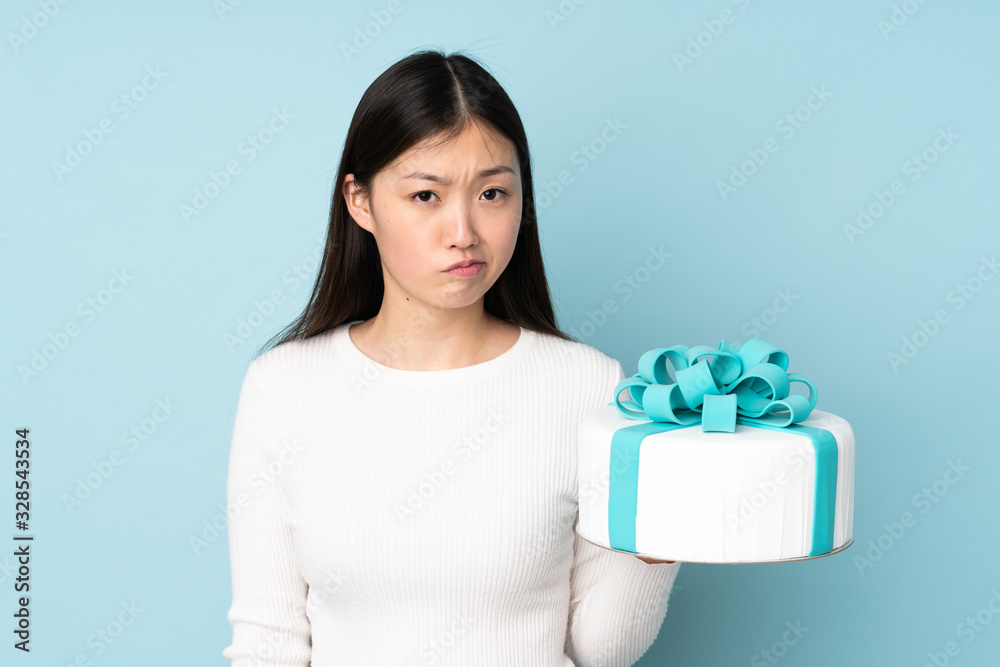 Pastry asian chef holding a big cake isolated on blue background with sad expression Pastry asian chef holding a big cake isolated on blue background with sad expression