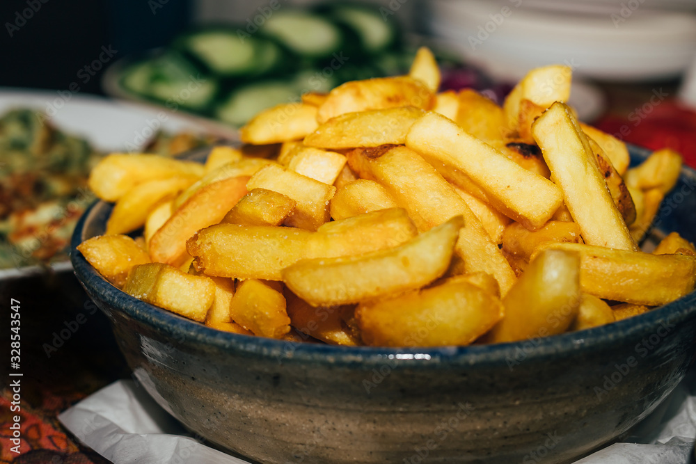 French fries served on a plate on table with blurred cucumbers on background, close-up. French fries served on a plate on table with blurred cucumbers on background, close-up.