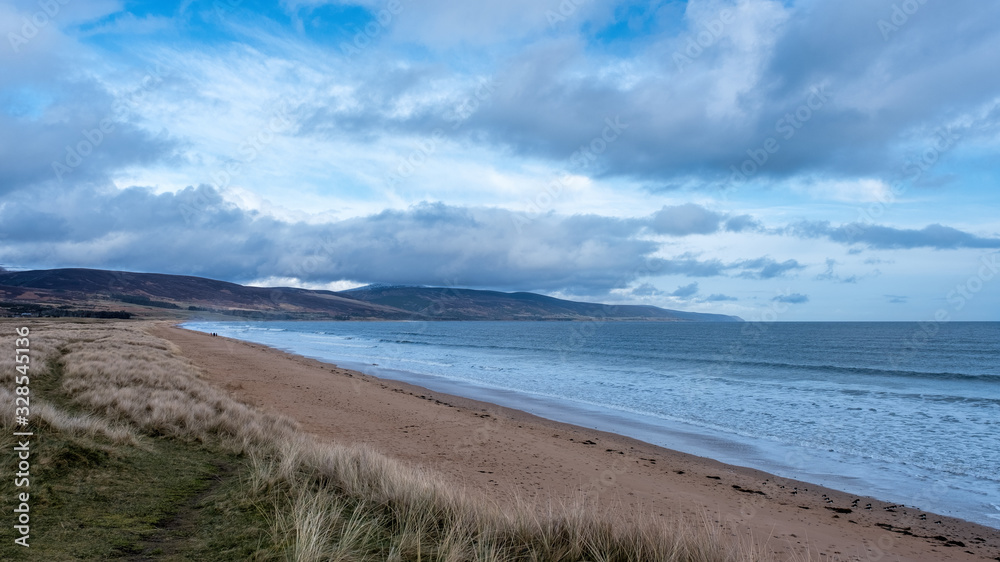Brora beach on a calm spring morning, with blue skies and clouds and with people walking the beach in the distance Brora beach on a calm spring morning, with blue skies and clouds and with people walking the beach in the distance
