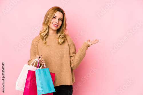 Young blonde woman holding shopping bags isolated showing a copy space on a palm and holding another hand on waist.