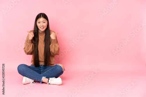 Young asian woman sitting on the floor isolated on pink background with thumbs up gesture and smiling