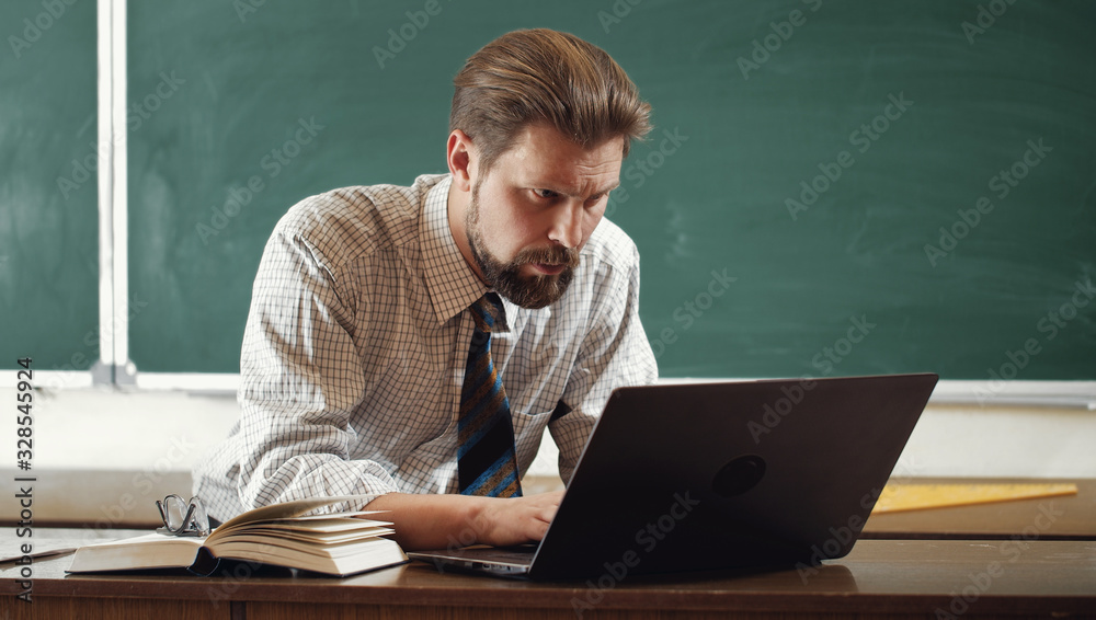 Portrait of absorbed adult bristled teacher sitting at desk and working on laptop in classroom
