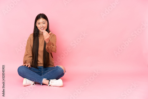 Young asian woman sitting on the floor isolated on pink background inviting to come with hand. Happy that you came