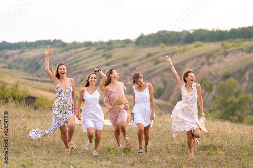 The company of cheerful female friends have a great time together on a picnic in a picturesque place overlooking the green hills. Girls in white dresses dancing in the field