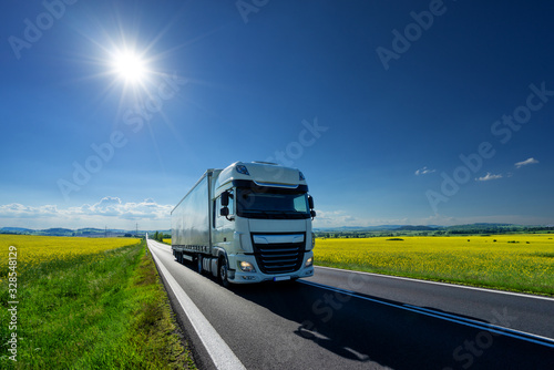 White truck driving on the asphalt road between the yellow flowering rapeseed fields under radiant sun in the rural landscape