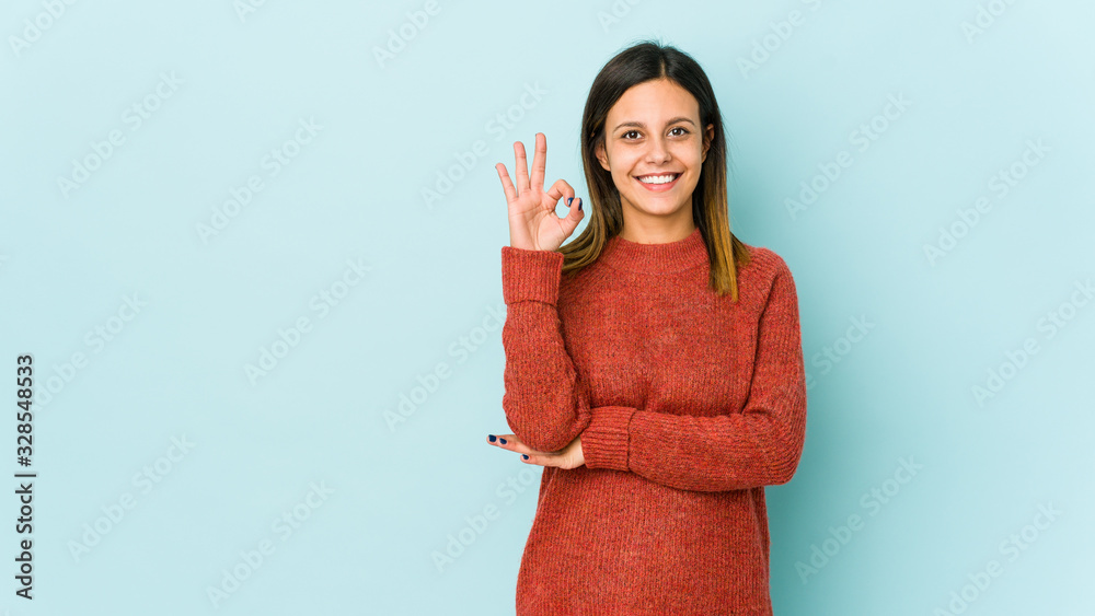 Young woman isolated on blue background winks an eye and holds an okay gesture with hand. Young woman isolated on blue background winks an eye and holds an okay gesture with hand.