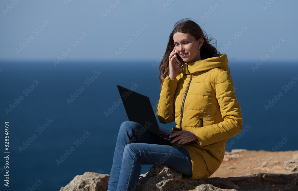 A freelancer girl is working typing on a laptop and talking on a mobile phone with a beautiful view of the open air sea sky. Traveling with a computer. Online dream job.