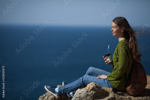 Beautiful young woman traveler with a backpack drinking morning coffee and looking at the sea. Copy space. The concept of freedom , morning coffee, travel and an active lifestyle.