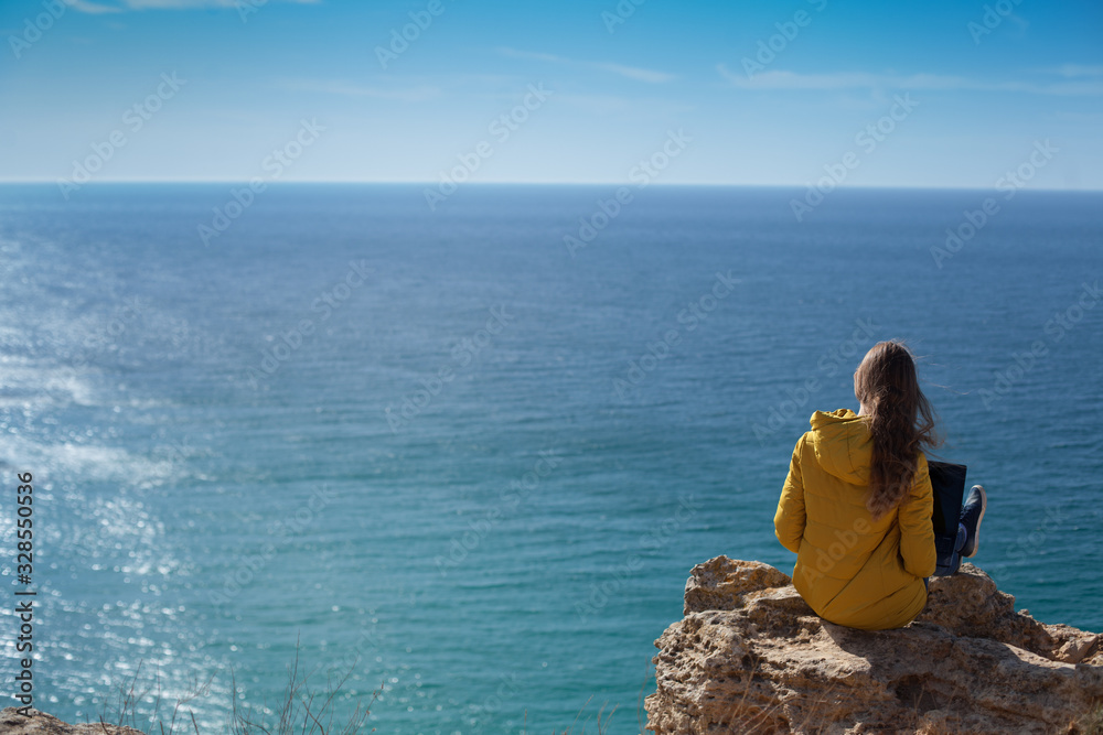 Technology and travel. Work in the open air. The concept of freelancing. A pretty young woman in a yellow jacket uses a laptop with a view of the sea Bay.