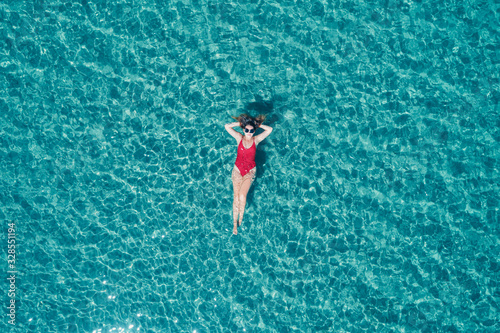 Top view of young pretty sexy girl in red swimsuit floating on water surface in crystal clear turquoise ocean. Happy island lifestyle.