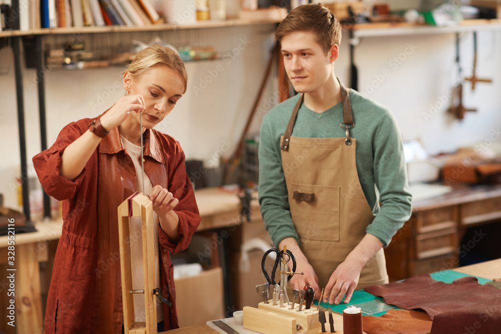 Horizontal medium portrait of female artisan using leather craft wooden clip and young man looking at her