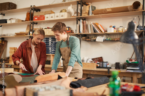 Attentive young man and concentrated woman creating new leather handicraft together in modern workshop