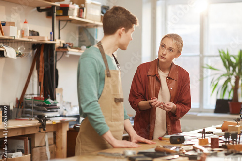 Horizontal shot of young man and woman working together in leather craft workshop standing at table discussing something