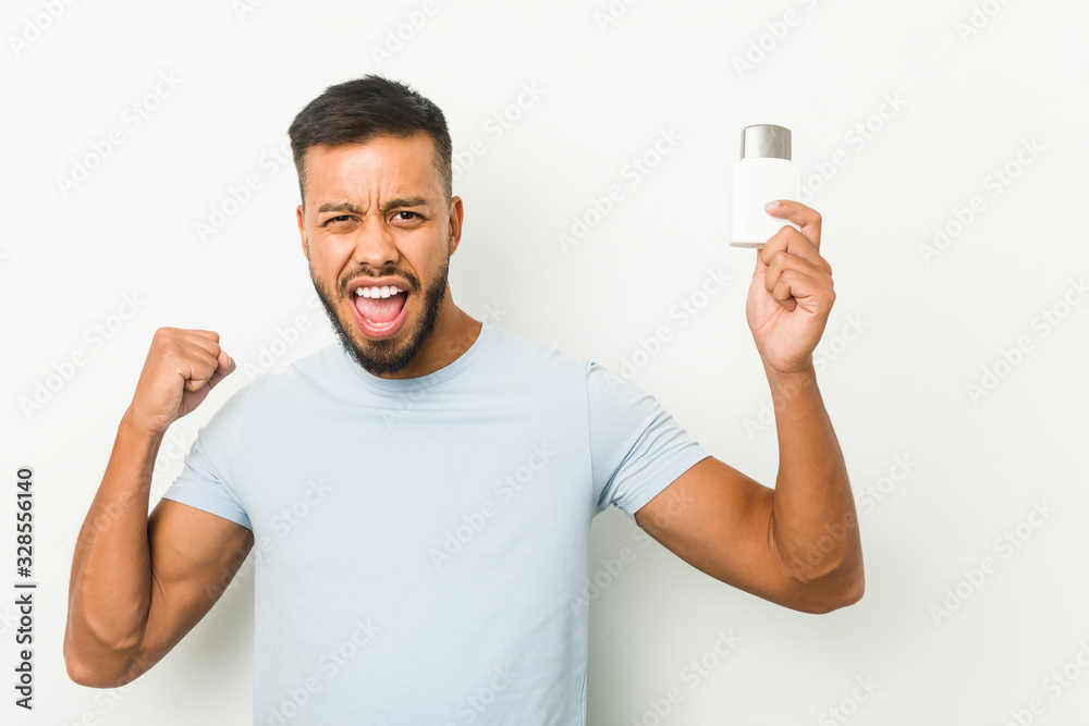 Young south-asian man holding an after shave cream raising fist after a victory, winner concept. Young south-asian man holding an after shave cream raising fist after a victory, winner concept.