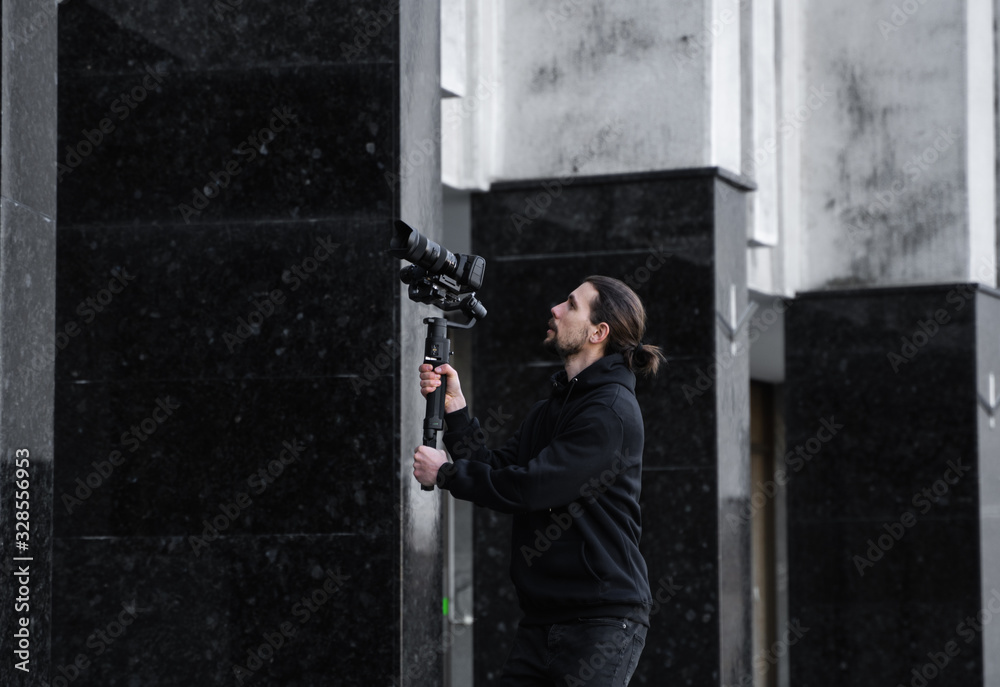 Young Professional videographer holding professional camera on 3-axis gimbal stabilizer. Pro equipment helps to make high quality video without shaking. Cameraman wearing black hoodie making a videos.