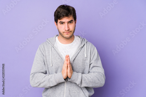 Young man isolated on purple background praying, showing devotion, religious person looking for divine inspiration. © Asier