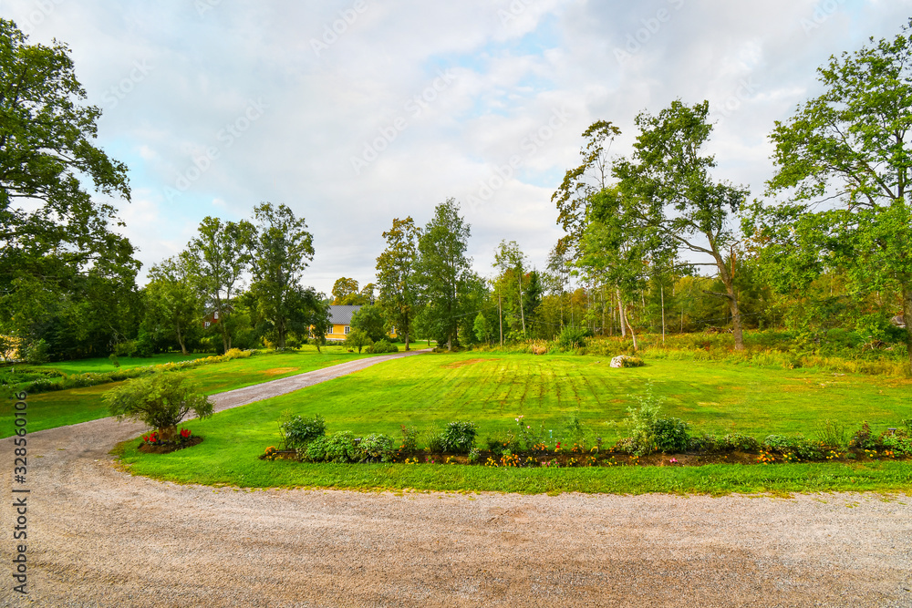A rural dirt road runs alongside a manicured garden and lawn near a yellow secluded home in the countryside of Finland.