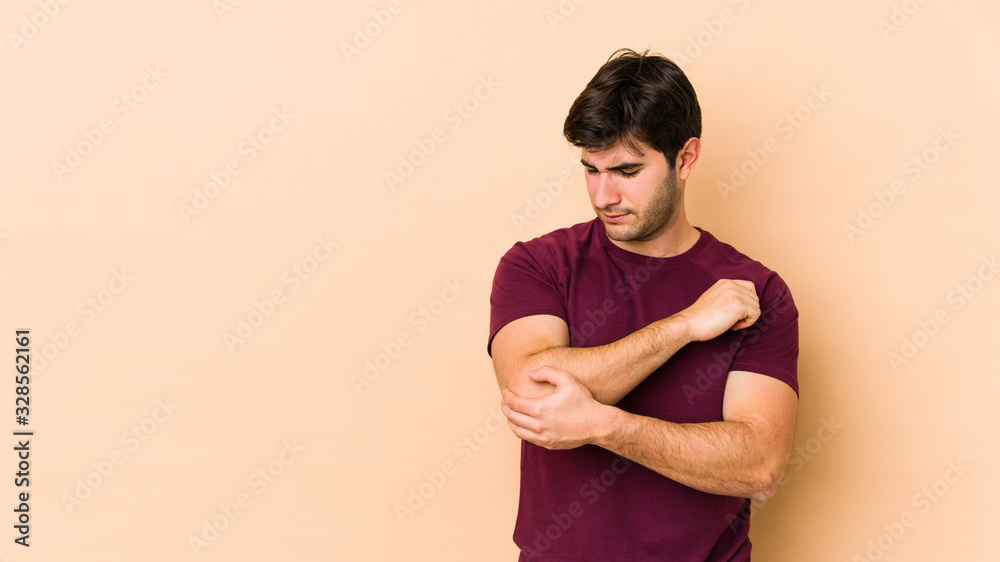 Young man isolated on beige background massaging elbow, suffering after a bad movement. Young man isolated on beige background massaging elbow, suffering after a bad movement.