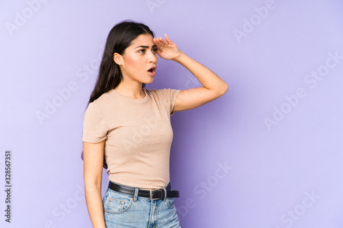 Young indian woman isolated on purple background looking far away keeping hand on forehead. © Asier