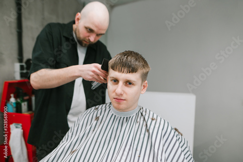 Handsome young man sits in a barber's chair, cuts a hairdresser's hair and looks into the camera with a serious face. Creating a stylish haircut in a barbershop. Attractive guy cares for hair