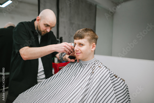 Portrait of a cheerful young man sitting in a peignoir in a barber shop chair and clipping at his hairdresser, looking at the camera and smiling.Professional barber cuts a positive young client