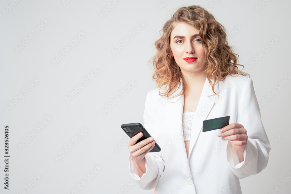 Serious curly blonde woman with red lips holding phone and credit card in her hands and looking at the camera, isolated over white background Serious curly blonde woman with red lips holding phone and credit card in her hands and looking at the camera, isolated over white background
