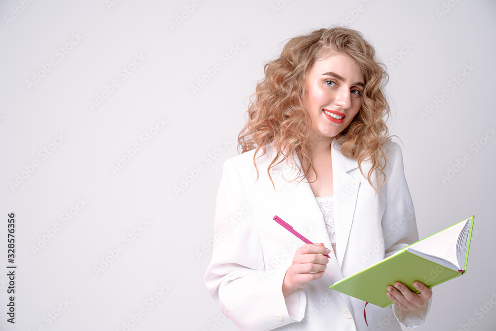 Attractive young woman with curly blonde hair and red lips holding green notebook and pink pen in her hands and looking at the camera Attractive young woman with curly blonde hair and red lips holding green notebook and pink pen in her hands and looking at the camera