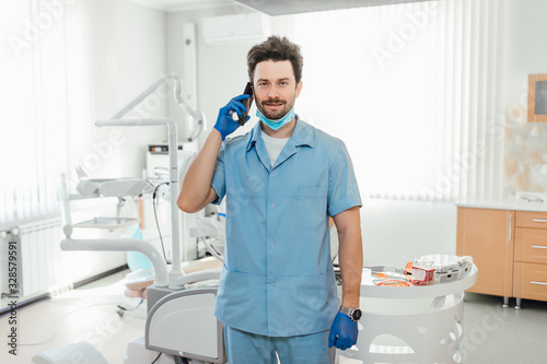 Healthcare, profession, stomatology and medicine concept - smiling male dentist over medical office background, holding phone.