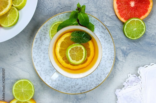 Slice of lemon and lime together with a basil leaf in a tea cup on an organte grey saucer on a grey textured surface surronded by citrus slices and a serviette photo