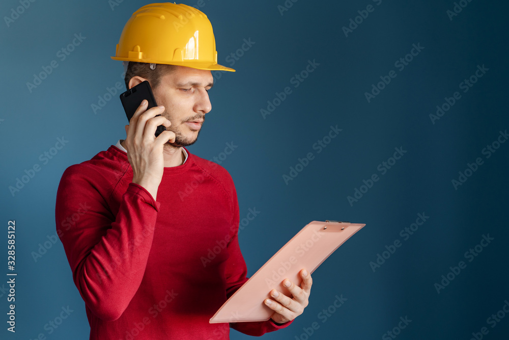 Portrait of young engineer or building contractor wearing yellow protective helmet and red sweater standing in front of blue wall background holding smart phone and clipboard call talking side view Portrait of young engineer or building contractor wearing yellow protective helmet and red sweater standing in front of blue wall background holding smart phone and clipboard call talking side view