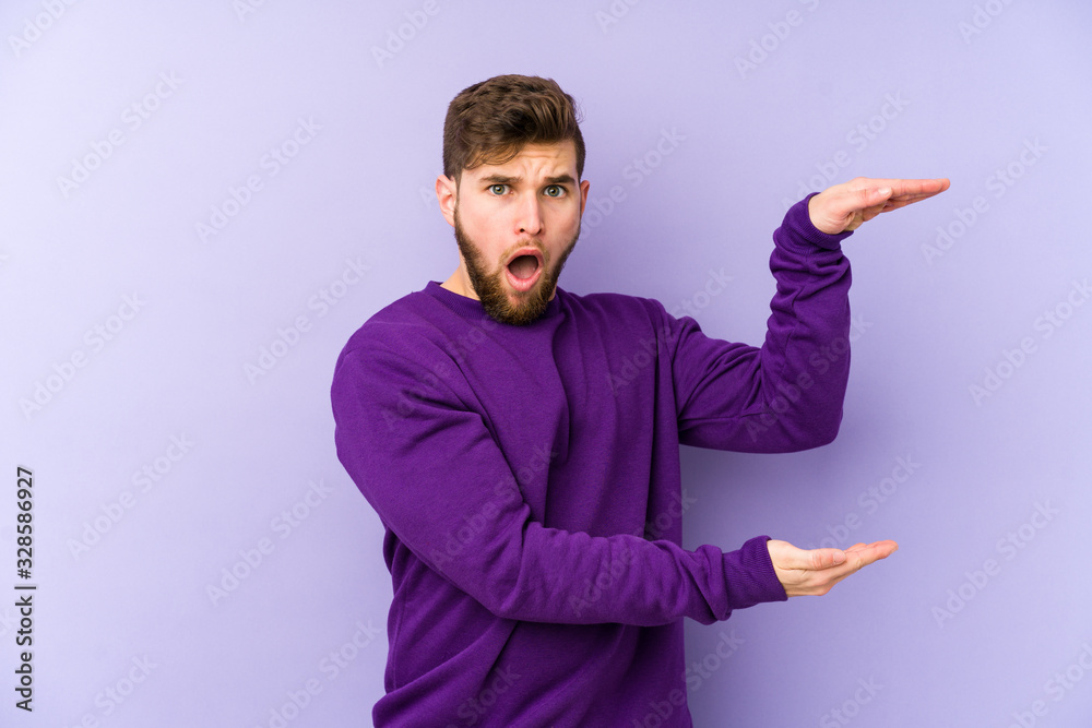 Young caucasian man isolated on purple background shocked and amazed holding a copy space between hands. Young caucasian man isolated on purple background shocked and amazed holding a copy space between hands.