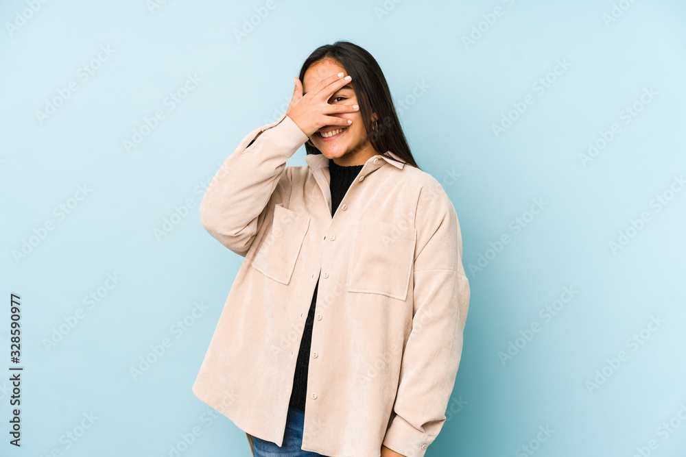 Young woman isolated on a blue background blink at the camera through fingers, embarrassed covering face. Young woman isolated on a blue background blink at the camera through fingers, embarrassed covering face.