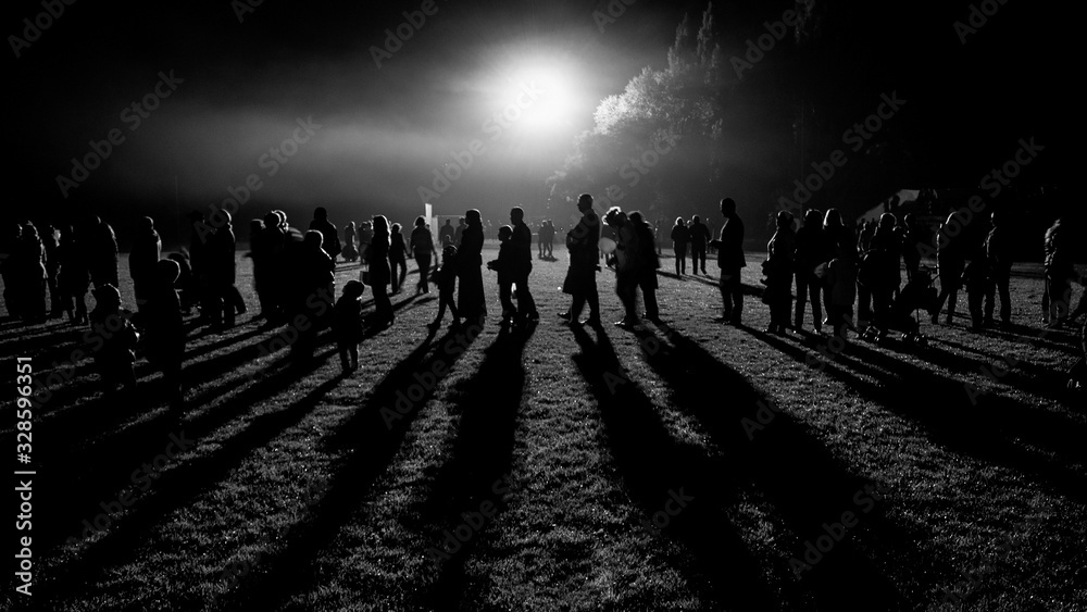 Black and white picture of a people standing one after another in front of bright light and casting long shadows on a grass on a local festival smoothed by a motion.