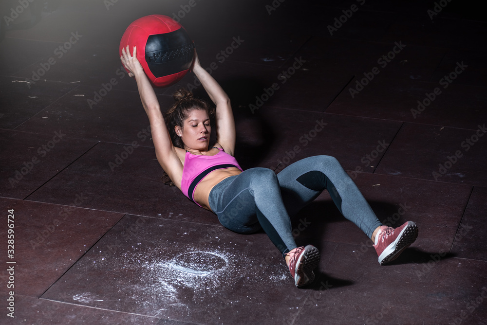 Young strong sweaty fit muscular girl with big muscles doing sit ups with medicine ball for abdominal muscles or abs hard crossfit workout training on the gym floor real people exercise