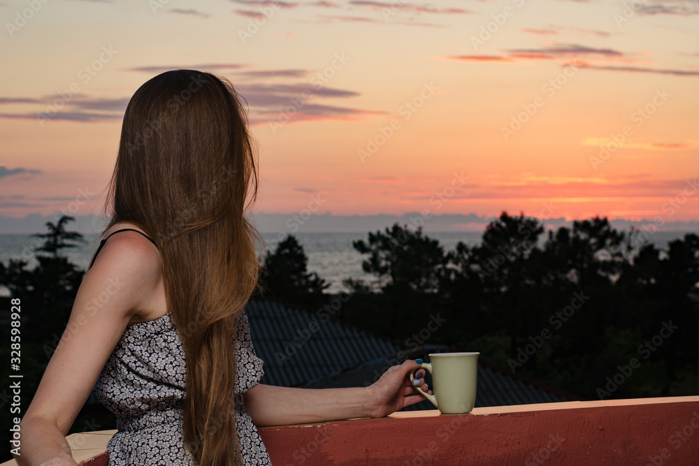 Fototapeta premium Young woman with a cup in her hand looking at sea and sunset. Side view