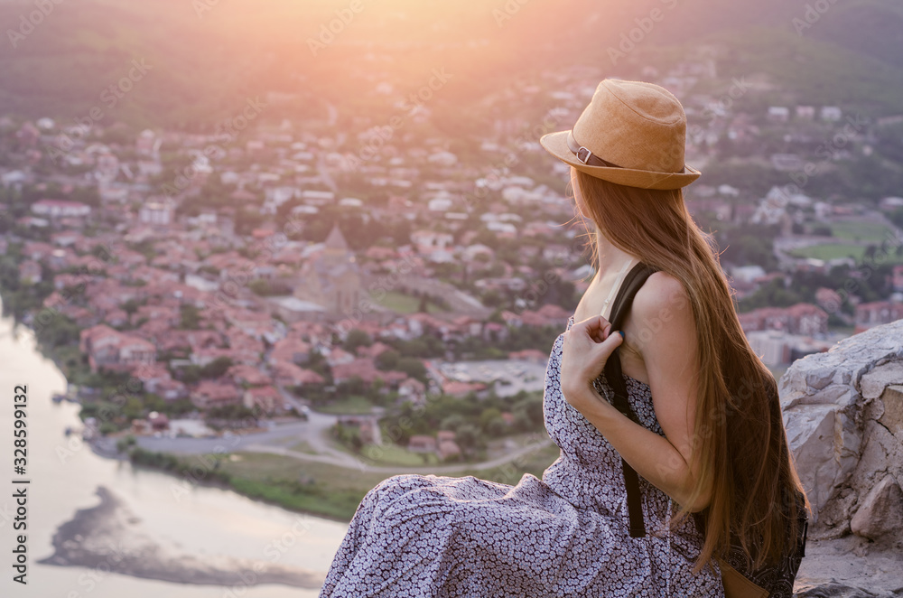 Young attractive girl sitting on hill looking at panorama of city in rays of sun. side view