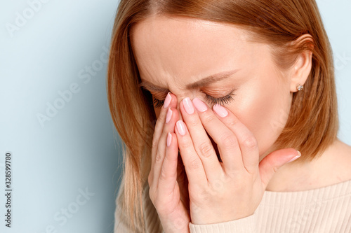 Close uo portrait of young woman has problem with contact lenses, rubbing her swollen eyes due to pollen, dust allergy. Dry eye syndrome, watery, itching, blue background.