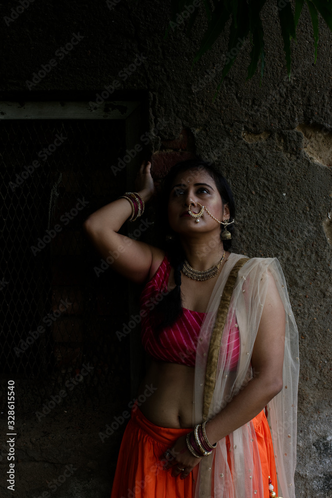 Fototapeta premium Young and attractive Indian Bengali brunette woman standing in front of a vintage house window wearing Indian traditional ethnic cloths pink and orange skirt blouse. Indian lifestyle and fashion