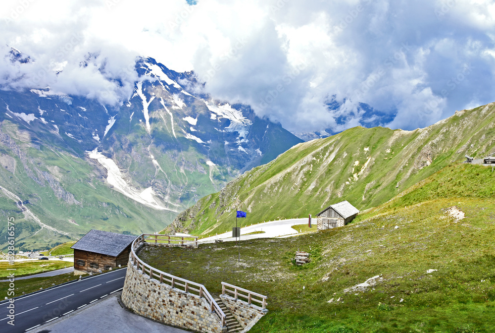 The Grossglockner High Alpine Road (in German Großglockner-Hochalpenstraße)  is the highest surfaced mountain pass road in Austria. wooden house with could and green mountain with snow in summer.