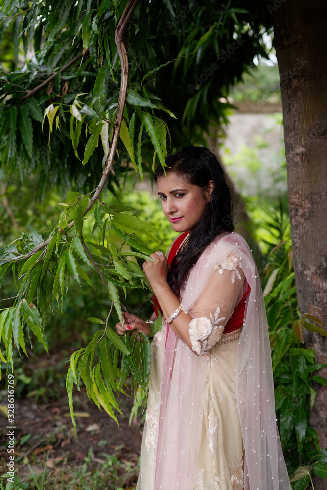 Young and beautiful Indian Bengali brunette woman standing thoughtfully in green nature below a tree wearing Indian traditional ethnic white and red gown. Indian lifestyle and fashion Young and beautiful Indian Bengali brunette woman standing thoughtfully in green nature below a tree wearing Indian traditional ethnic white and red gown. Indian lifestyle and fashion