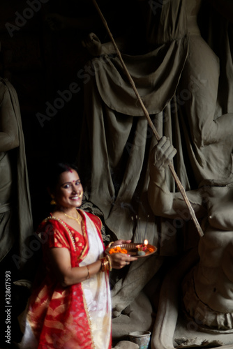 An young and beautiful Indian Bengali brunette woman in red white traditional ethnic sari holding a puja thali for worshiping goddess Durga clay idol . Indian culture, religion and fashion